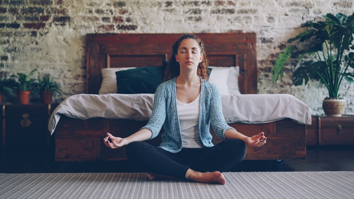 Beautiful young lady is sitting in lotus position on bedroom floor and enjoying meditation, harmony and peace. Modern loft style houses, healthy people and rest concept.
